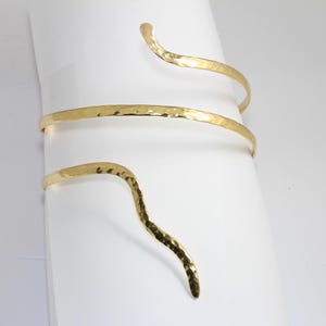 May include: A gold hammered metal cuff bracelet with a wavy design. The bracelet is made of three separate pieces of metal that are connected together.