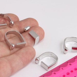 Könnte beinhalten: Vier silberne Ringe mit abgerundeter Kante und einer flachen rechteckigen Leiste. Die Ringe befinden sich auf einer Hand und einem rosa Lineal.