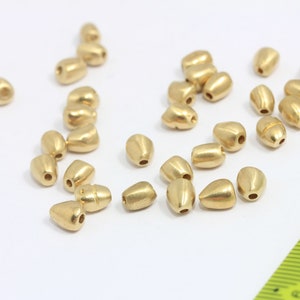 Könnte beinhalten: Eine Gruppe von 25 goldfarbenen Metallperlen, jede in Tropfenform mit einem kleinen Loch oben. Die Perlen sind auf einer weißen Oberfläche verstreut.