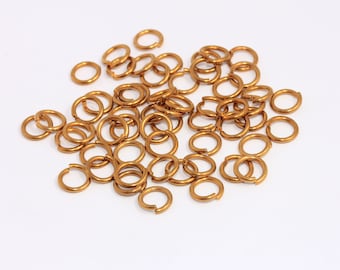 Puede incluir: Un conjunto de pequeños anillos abiertos de color oro metálico cálido. Estos anillos circulares están dispersos sobre una superficie blanca, ideales para la fabricación de joyas y proyectos de artesanía. Los anillos son de tamaño y forma uniformes.