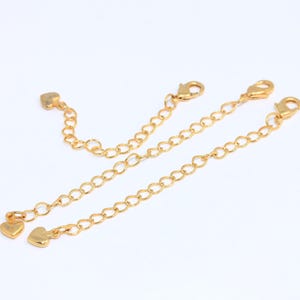 May include: Gold-colored necklace extenders with lobster claw clasps and heart-shaped end pieces. The extenders are of varying lengths, with a small heart charm on one end. The chains are on a white background.