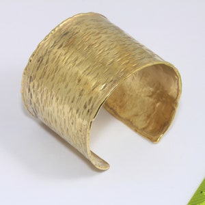 Op de afbeelding: Een gouden metalen armband met een gehammerde textuur. De armband is open en heeft een breed, plat ontwerp.