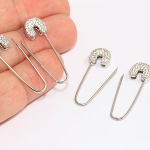 Könnte beinhalten: Vier silberne Sicherheitsnadel-Ohrringe mit klaren Strasssteinen.