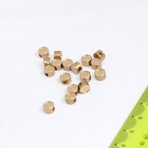 Può includere: Un mucchio di piccole perline di metallo dorate con un foro al centro. Le perline sono sparse su una superficie bianca accanto a un righello verde.