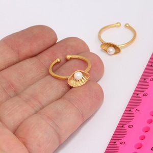 May include: Two gold rings with a pearl and a shell design. The rings are on a hand against a white background.