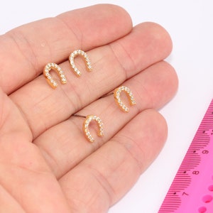 May include: Three pairs of gold horseshoe earrings with clear cubic zirconia stones. The earrings are shown on a hand with a pink ruler for size reference.