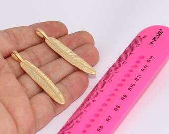 May include: Two gold-colored feather charms, each approximately 3.5 inches long, are shown on a hand against a pink ruler. The ruler has markings in inches and centimeters.