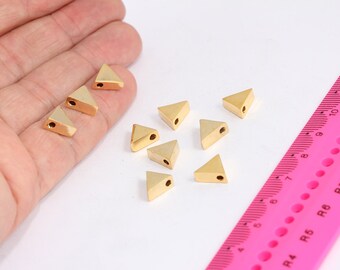 May include: A collection of ten gold-colored triangle-shaped metal beads with a hole in the center. The beads are scattered on a white surface.