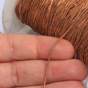 May include: Close-up of a spool of copper-colored cord. A strand of the cord is held between fingers, showcasing its fine, flexible texture. The cord is tightly wound on a white spool, suggesting its use in crafts or jewelry making.