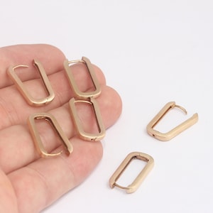 Puede incluir: Cinco pendientes de aro rectangulares de color dorado. Los pendientes están en una mano y una superficie blanca.