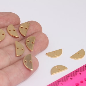May include: A close-up of eight gold-colored metal half-circle shapes with two holes each. The shapes are arranged on a hand and a pink ruler.