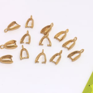 Puede incluir: Una colección de pequeños hallazgos de joyería dorados, probablemente conectores de fianza, dispuestos sobre una superficie blanca. Cada pieza tiene un lazo en la parte superior y una abertura en forma de U en la parte inferior. Una regla amarilla está en el fondo.