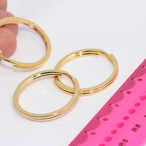 Può includere: Tre portachiavi dorati, ognuno con un design a doppio anello, sono disposti su una superficie bianca. Un righello rosa con segni neri è parzialmente visibile sullo sfondo.