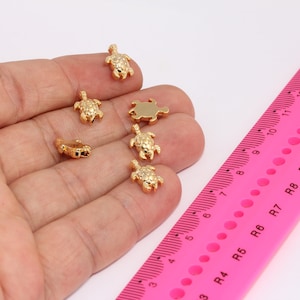 May include: Six gold-coloured turtle charms, some with textured shells, are displayed on a hand against a pink ruler with black markings.