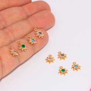 May include: Six gold-tone star-shaped charms with different colored rhinestones. The charms are on a white surface and a pink ruler is in the background.