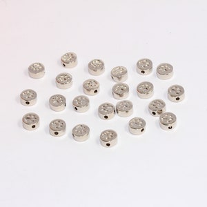 May include: A collection of small, silver-colored metal beads. Each bead is round with a flat top featuring a decorative pattern and a small hole for stringing. The beads are scattered on a white surface.