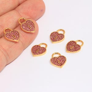 May include: Five gold heart-shaped charms with a keyhole design. Each charm is covered in pink rhinestones.