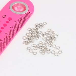 Op de afbeelding: Een roze plastic liniaal met een heldere vergrootglas en genummerde gaten met de labels R2 tot en met R8. Een stapel zilveren springringen ligt verspreid op een wit oppervlak.