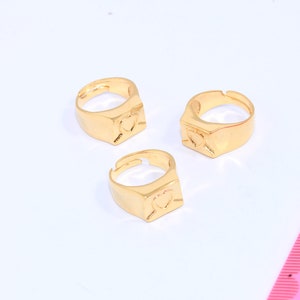 以下が含まれることがあります： ハート型のデザインが施された金色の指輪が3つ。指輪は白い表面の上に置かれています。