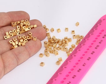 May include: A large pile of small, gold-colored, square-shaped beads with a hole in the center. The beads are scattered on a white surface next to a pink ruler.