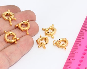 Puede incluir: Seis cierres de metal de color dorado, cada uno con un anillo circular y dos pequeños bucles, están dispuestos en una mano y una regla rosa. La regla está marcada en pulgadas y centímetros.