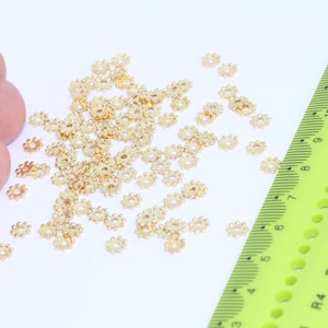 May include: A large quantity of small, gold-colored metal beads with a starburst design. The beads are scattered on a white surface next to a green ruler.