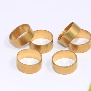 Puede incluir: Seis anillos de metal de color dorado, probablemente para la fabricación de joyas o proyectos de artesanía, están dispuestos sobre una superficie blanca. Una regla verde es visible en el fondo.