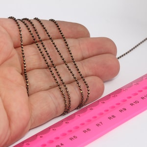 May include: A close-up of a bronze-colored chain with small round beads. The chain is laid out on a pink ruler with markings for ring sizes. The ruler is labeled "V-PLUS" and has markings for sizes 6 through 13.