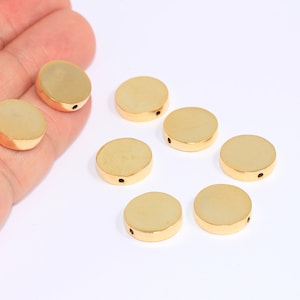 May include: Eight gold-coloured round metal beads with a small hole in the centre. The beads are arranged in a circle on a white surface.