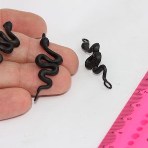 Puede incluir: Tres abalorios negros en forma de serpiente con un bucle en el extremo. Los abalorios miden aproximadamente 5 cm de largo y se muestran en una mano junto a una regla rosa.