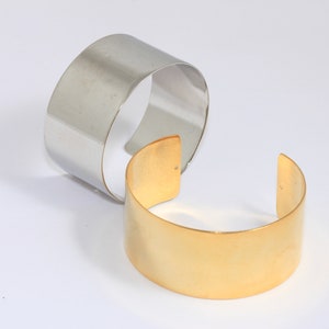 May include: Two metal bracelets, one silver and one gold, are stacked on top of each other. The bracelets are both wide and flat.