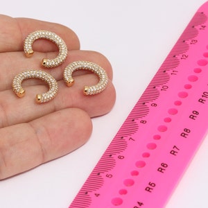 May include: Three gold-colored, curved ear cuffs encrusted with small, clear crystals. The ear cuffs are arranged on a light-colored surface. A pink ruler with centimeter markings is in the background.