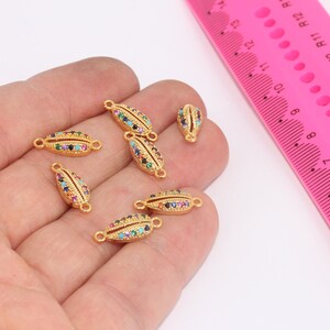 Könnte beinhalten: Nahaufnahme von sechs goldfarbenen muschelförmigen Verbindungsanhängern mit bunten Strasssteinen. Die Anhänger liegen auf einer Hand, im Hintergrund befindet sich ein rosa Lineal.