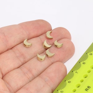May include: Six gold-colored crescent moon-shaped beads, approximately 1/2 inch in diameter, are displayed in a hand. The beads have a small hole in the center for stringing.