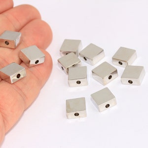 Peut inclure: Un groupe de perles carrées en métal argenté avec un trou au centre. Les perles sont éparpillées sur une surface blanche.