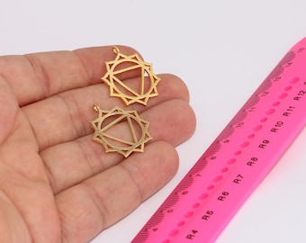 May include: Two gold-colored metal charms in the shape of a mandala with a triangle in the center. The charms are on a hand and a pink ruler with black markings.