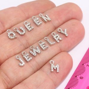 Puede incluir: Primer plano de una mano sosteniendo un conjunto de colgantes de plata con las letras "QUEEN" y "JEWELRY" escritas en pedrería. La letra "M" también está incluida en el conjunto.