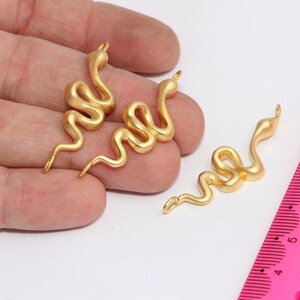 Può includere: Tre ciondoli a forma di serpente color oro con finitura opaca. I ciondoli sono a forma di spirale, con un anello nella parte superiore per appenderli. Un righello rosa è sullo sfondo.