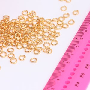 以下が含まれることがあります： 白い表面に散らばったたくさんの金色のジャンプリングと、黒い目盛りが付いたピンクの定規。