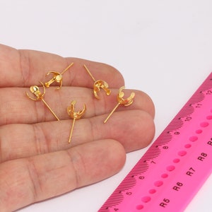May include: Five gold-colored earring settings with prongs, each with a small, round, gold-colored post. The settings are shown on a hand, next to a pink ruler with black markings.
