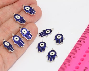 May include: A collection of blue and silver hamsa hand charms with an evil eye design. The charms are arranged on a hand and a pink ruler.