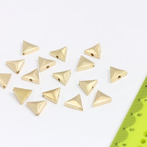 Peut inclure: Un ensemble de 18 perles en métal de forme triangulaire de couleur dorée avec un trou au centre. Les perles sont éparpillées sur une surface blanche à côté d'une règle verte.