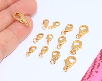 Puede incluir: Una colección de cierres de langosta dorados, comúnmente utilizados en la fabricación de joyas. Los cierres están dispuestos sobre una superficie blanca, con una regla rosa visible en el fondo. Los cierres son pequeños, con un diseño curvo y un mecanismo de resorte.