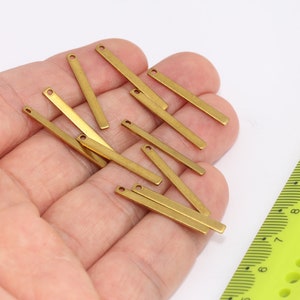 May include: A selection of small, rectangular brass bars, each with a single hole at one end. The bars are held in a hand, with a yellow ruler in the background. These components are likely for jewellery making or other craft projects.