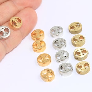 Peut inclure: Un ensemble de petites perles rondes en métal doré et argenté. Chaque perle présente un motif découpé et de petits trous. Les perles sont disposées sur une surface blanche, certaines étant tenues dans une main.