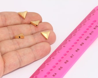 May include: Four gold-colored triangular metal pyramid shapes with a hole in the center. The pyramids are on a hand, and a pink ruler with black markings is in the background.