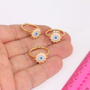 May include: Three gold rings with white and blue evil eye charms. The rings are on a hand against a white background.