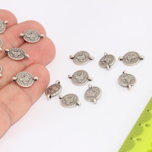 Può includere: Una collezione di ciondoli in metallo color argento con un design decorativo. I ciondoli hanno un anello su ogni lato per essere attaccati a gioielli o altri oggetti artigianali.