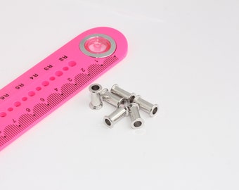 May include: A close-up of six silver metal grommets, a common hardware item used for reinforcing holes in fabric, leather, or other materials. The grommets are arranged in a loose pile on a white surface. A pink ruler with black markings is visible in the background.