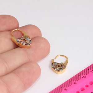 Peut inclure: Une paire de boucles d'oreilles en forme de cadenas en or avec des pierres précieuses colorées. Les boucles d'oreilles sont présentées sur une main et à côté d'une règle rose.